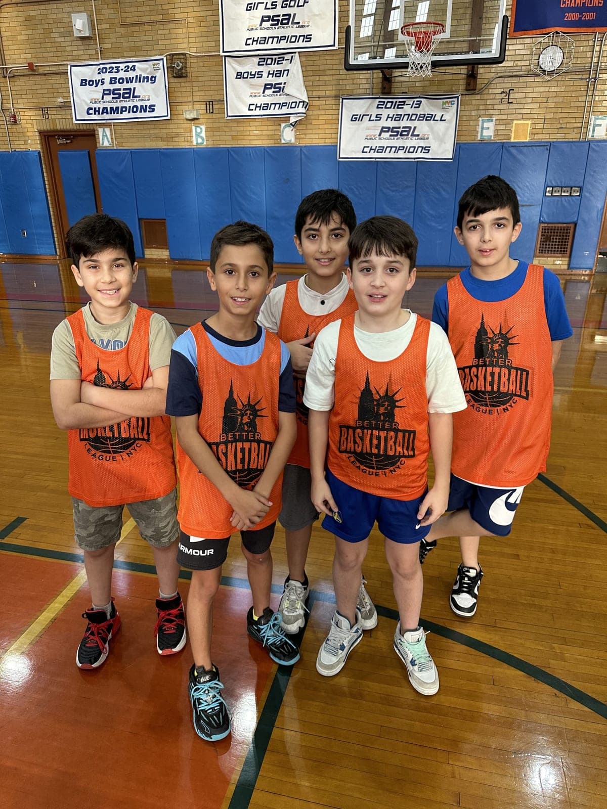 Five players in orange BBL NYC pinnies posing under the hoop, championship banners on the wall behind them