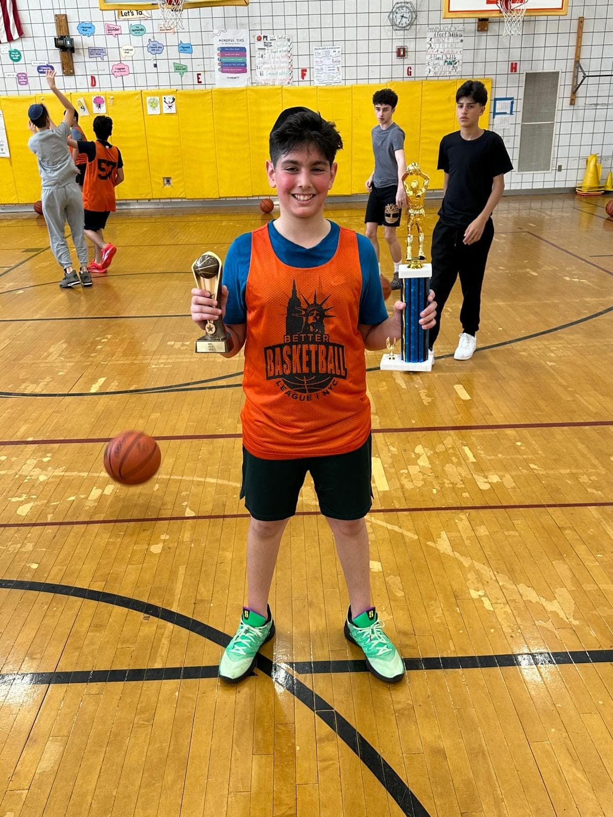 A player in an orange pinnie holding an end-of-season award, a taller trophy standing on the floor beside him while a shootaround goes on behind