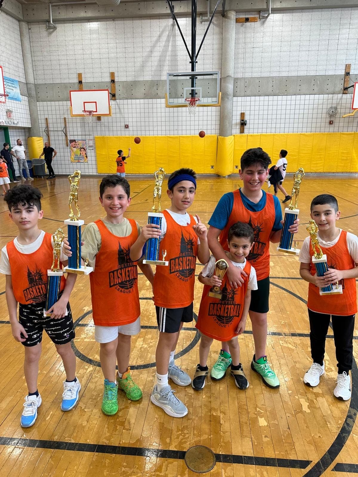 Six players in orange BBL NYC pinnies at center court, each holding a tall championship trophy