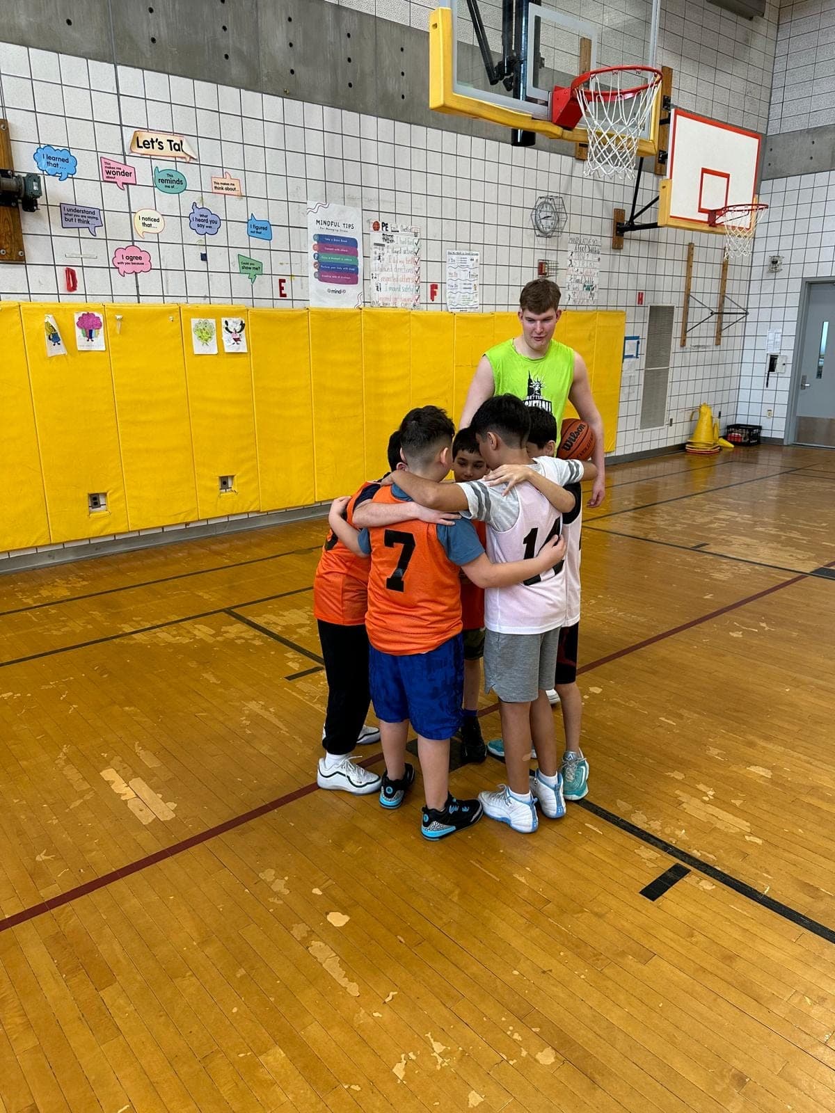 Five players locked arm in arm in a huddle, a coach standing with them holding the ball