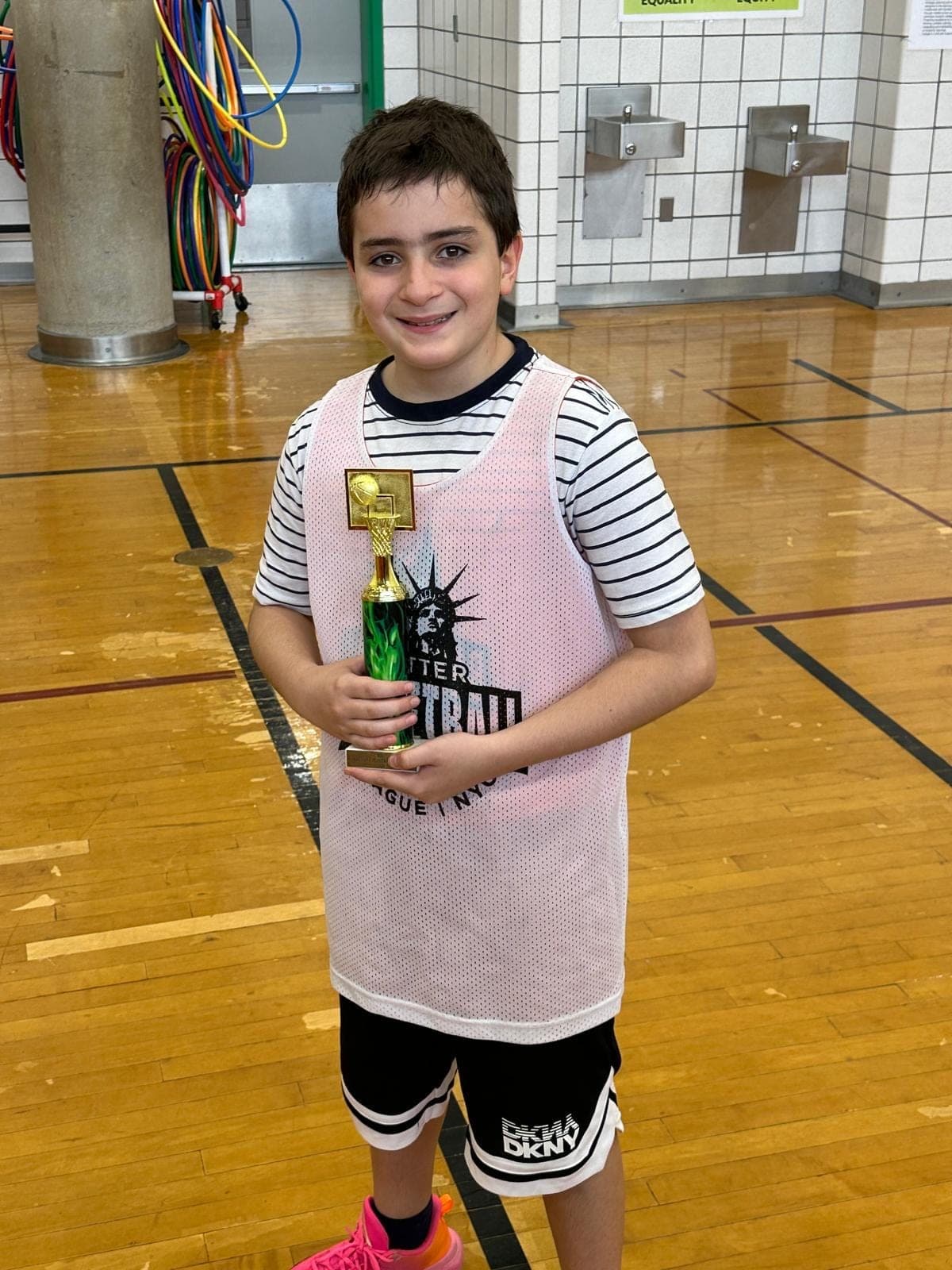 A player in a white BBL NYC pinnie holding his end-of-season trophy at the side of the gym